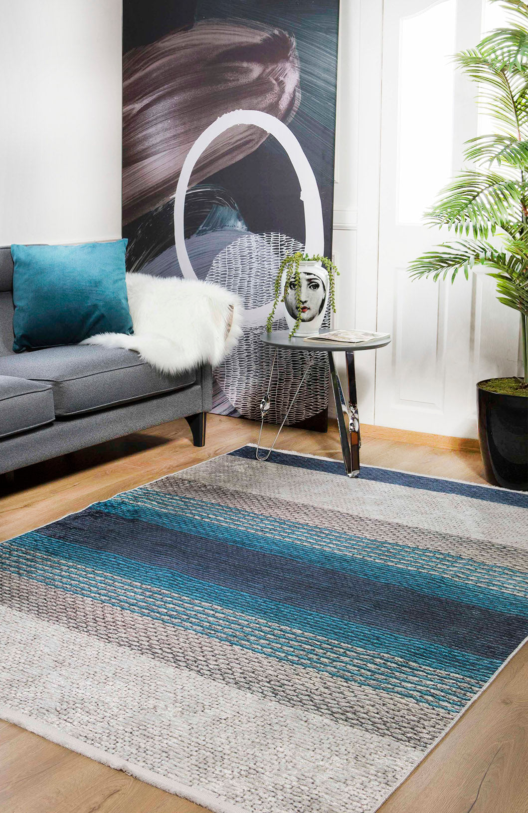 A modern living room with a gray sofa, teal pillow, white throw, abstract wall art, small glass table with face planter, large green plant, and the Artistic Angles - Washable Rug - JR908—ideal for high-traffic areas—on wood floors.