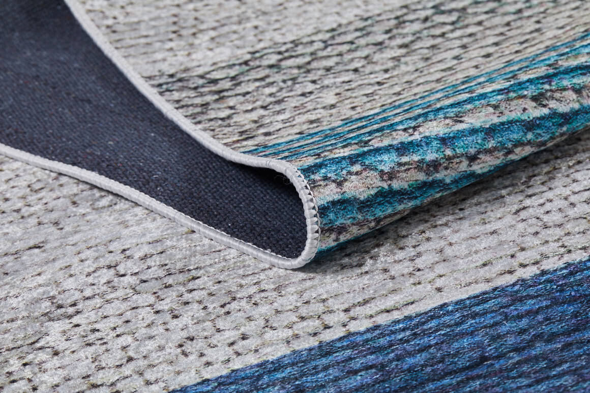 A close-up of the Artistic Angles - Washable Rug - JR908 highlights its textured blue, gray, and teal shades. Slightly folded to reveal thickness and detailed edge stitching, this rug is ideal for high-traffic areas.