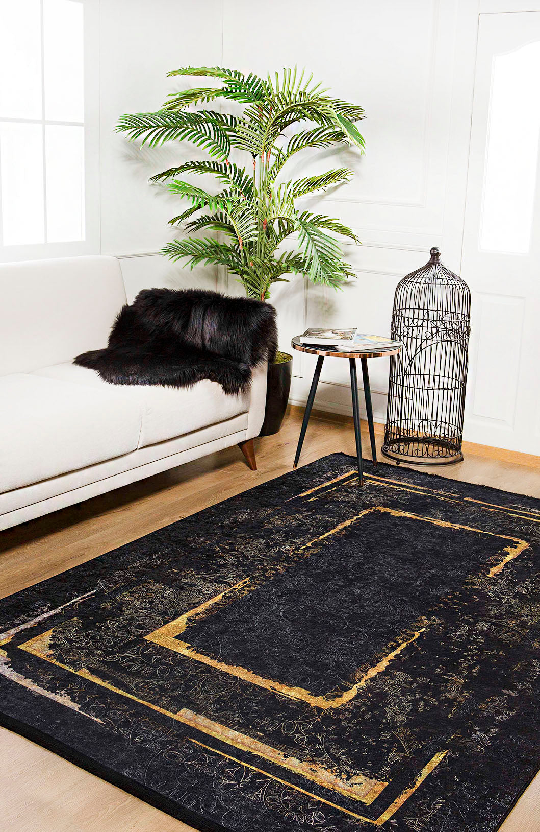 A modern living room features a white sofa, black fur throw, round side table, birdcage, potted palm, and the Azure Glow - Washable Rug - JR1337 with gold geometric accents—ideal for high-traffic areas. Sunlight pours in from large windows.