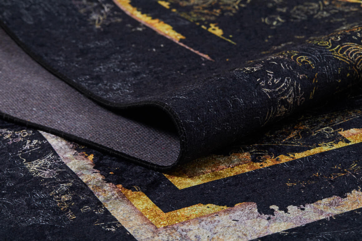 A close-up of the Azure Glow - Washable Rug - JR1337 shows its dark, textured black surface with gold and yellow abstract patterns, slightly folded to reveal layered colors—perfect for high-traffic areas.