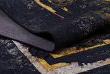 A close-up of the Azure Glow - Washable Rug - JR1337 shows its dark, textured black surface with gold and yellow abstract patterns, slightly folded to reveal layered colors—perfect for high-traffic areas.