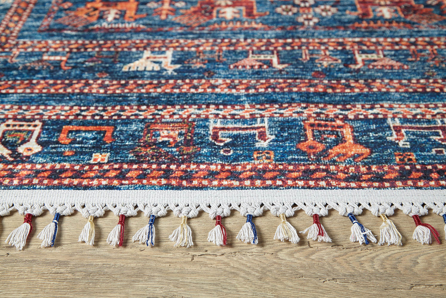 A close-up of the Berber Beauty - Washable Rug - JR5092, featuring blue with red and cream geometric patterns and colorful tassels on the edge, placed on a wooden floor—ideal for high-traffic areas.