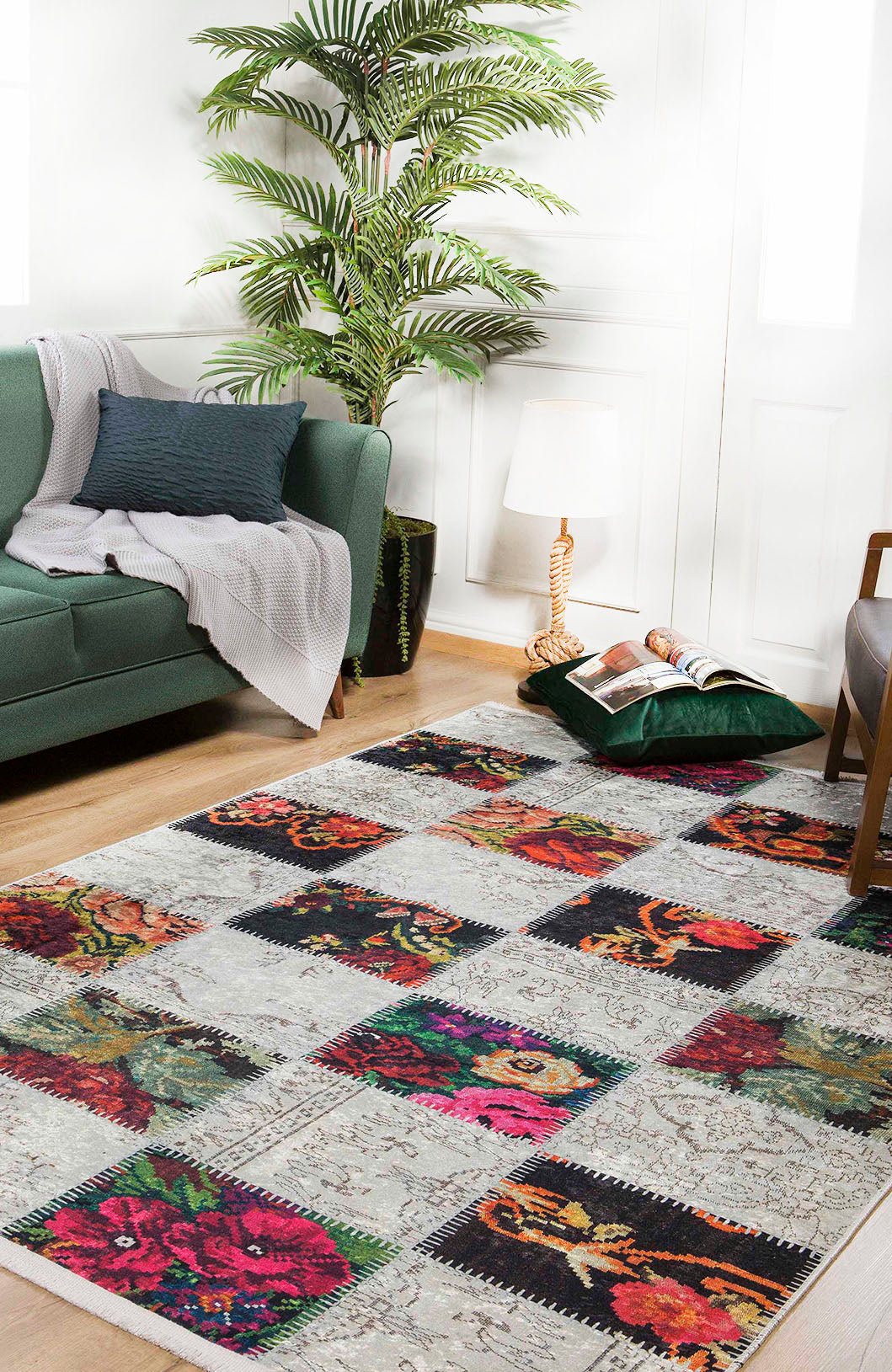 A cozy living room with a green sofa, gray blanket, pillow, large potted plant, floor lamp, magazines, and the Blooming Paradise - Washable Rug - JR188—a multicolor floral rug ideal for high-traffic areas on light wooden floors.