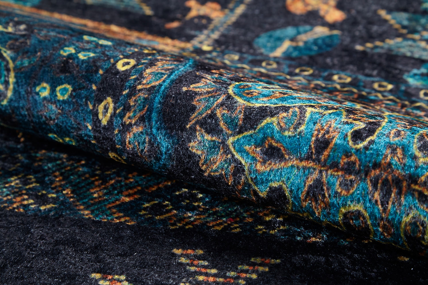 Close-up of the Blossoming Bazaar - Washable Rug (JR1862): a rolled-up, dark-colored rug featuring intricate blue, teal, and yellow floral patterns on black. Its visible texture and durable weave make it ideal for high-traffic areas.