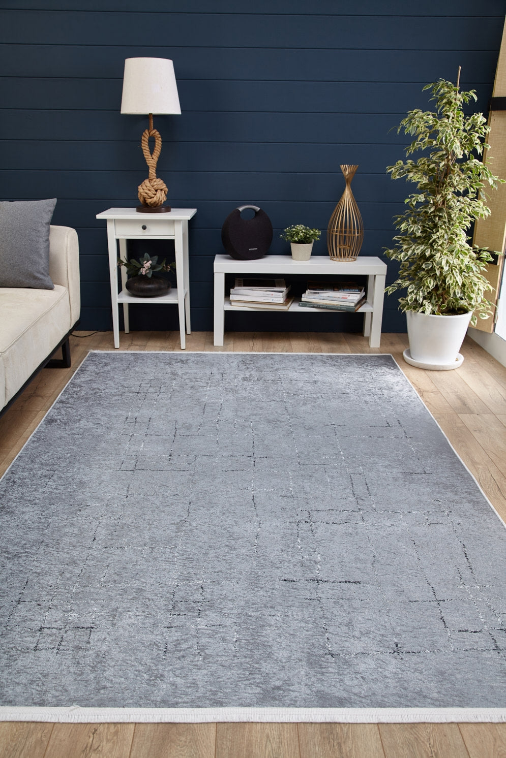A modern living room featuring the Bohemian Driftwood - Washable Rug - JR1951 in gray-anthracite on wood floors, paired with a white sofa, two white tables with decor, a lamp, books, and a potted plant against dark blue paneled walls.