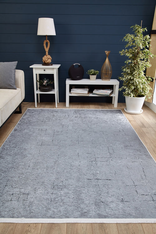 Load image 1 in gallery view. A modern living room featuring the Bohemian Driftwood - Washable Rug - JR1951 in gray-anthracite on wood floors, paired with a white sofa, two white tables with decor, a lamp, books, and a potted plant against dark blue paneled walls.