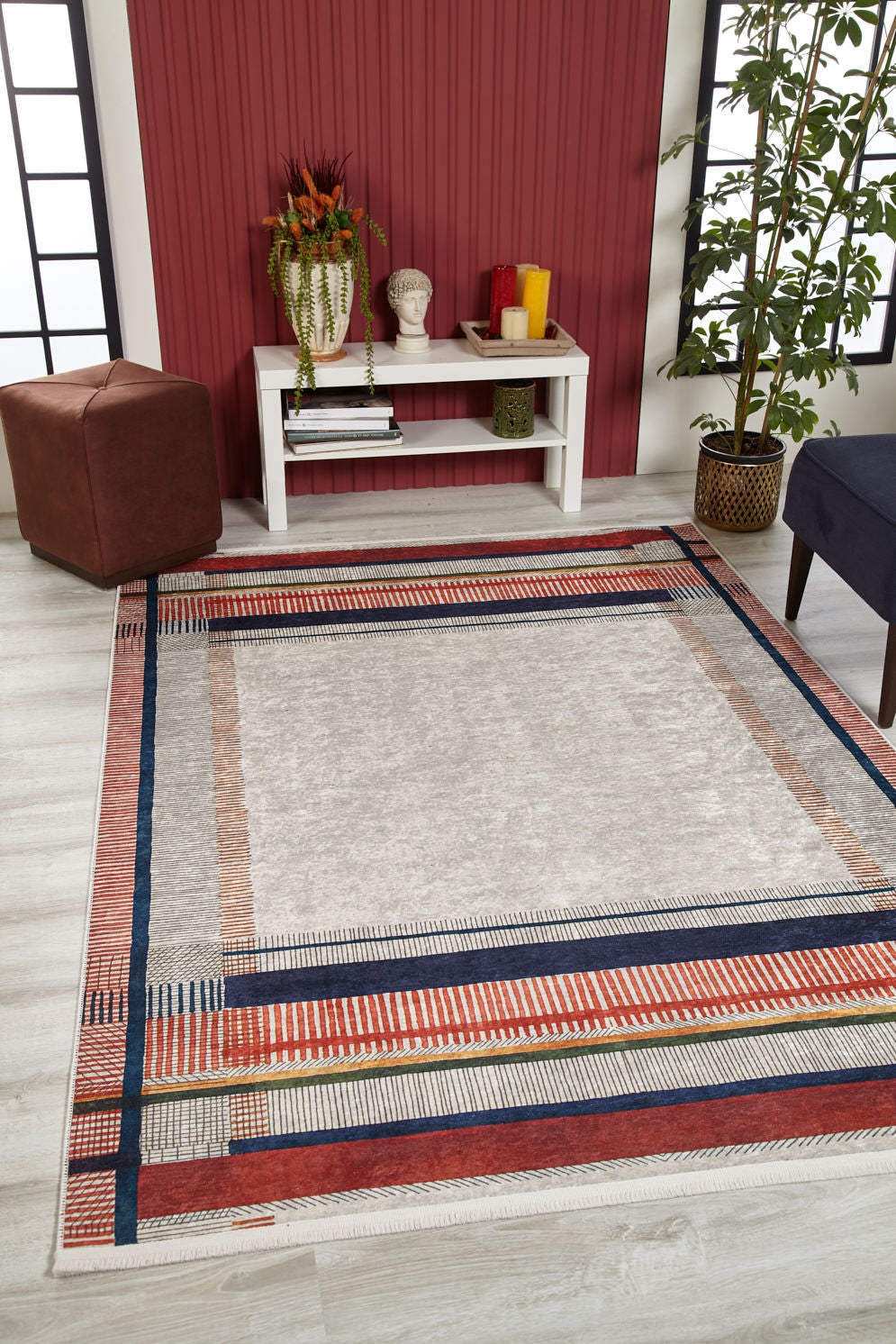 A modern living room with the Bohemian Nightscape - Washable Rug - JR1701, featuring red, navy, and beige stripes—ideal for high-traffic areas—light wood floor, white console table, brown ottoman, blue chair, and plant by a red accent wall.