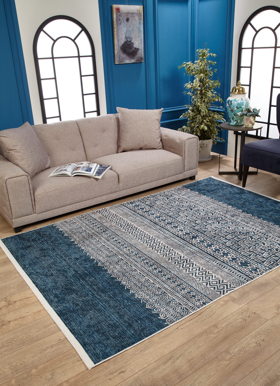A modern living room with a beige sofa, blue-paneled walls, and the Bohemian Retreat - Washable Rug - JR1589. A navy chair, plant, small side table with a lamp, and decor complete the space. Light streams in through arched windows—ideal for busy areas.