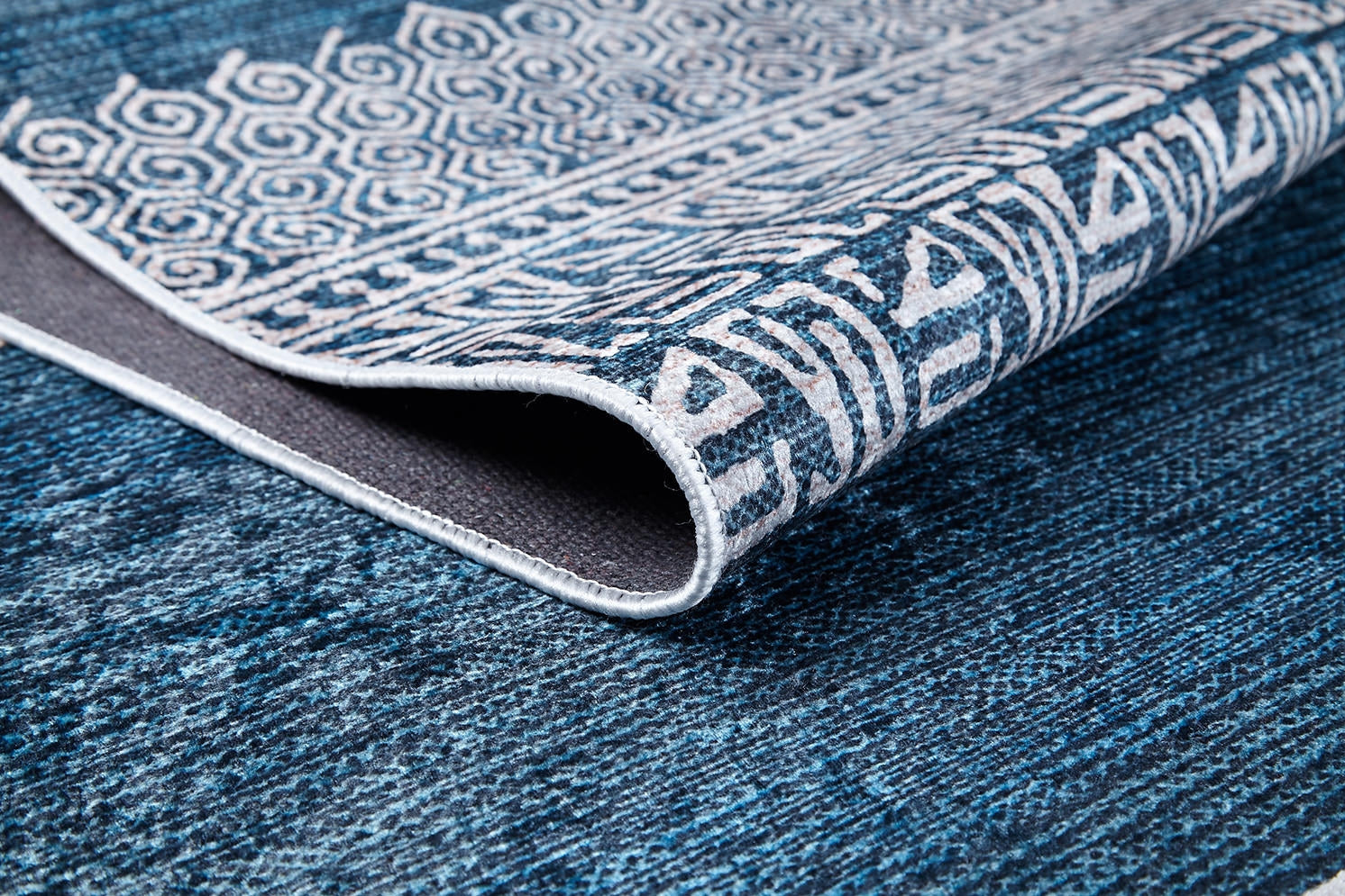A close-up of the Bohemian Retreat - Washable Rug - JR1589 shows blue tones with white geometric and abstract patterns, a folded corner revealing its texture—ideal as a washable rug for high-traffic areas.