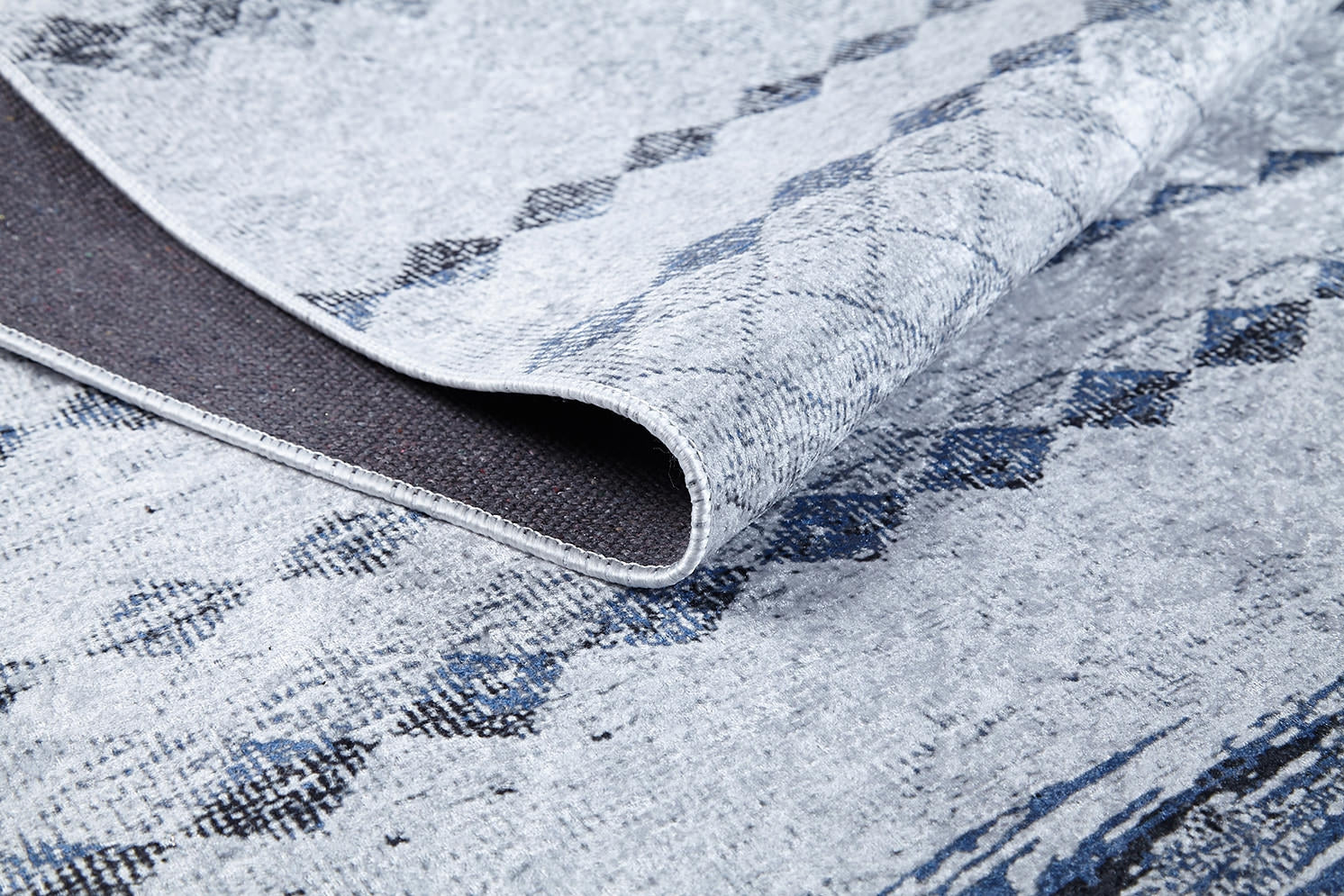 A close-up of the Bohemian Reverie - Washable Rug - JR1974 (Outlet) US shows its light gray and blue pattern, a folded corner revealing the dark, textured backing and detailed weave—ideal for high-traffic areas.