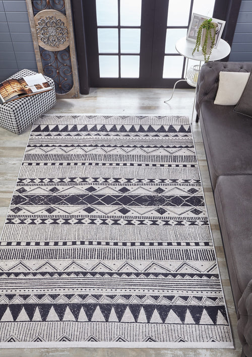 Load image 1 in gallery view. A modern living room with the Bohemian Sunrise - Washable Rug - JR1713 in gray-anthracite, featuring a black-and-white geometric pattern, gray sofa, side table with plant, wall art, and pouf—perfect for high-traffic areas.