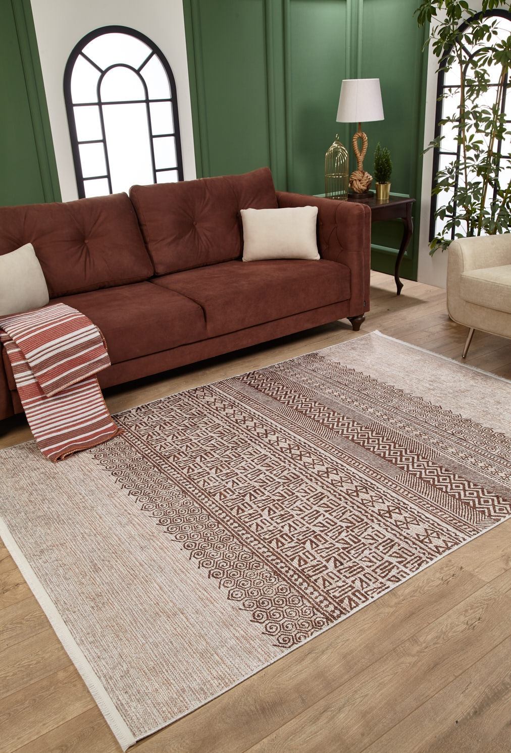 A living room featuring a brown sofa, white cushion, striped throw blanket, Bohemian Treasure - Washable Rug - JR1586—ideal for high-traffic areas—a lamp on a side table, green paneled walls with arched windows, beige chair, and plant.