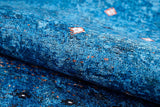 A close-up of the Bohemian Whirlwind - Washable Rug (JR1989) in blue, featuring red, white, and black square patterns on its textured, washable surface—ideal for high-traffic areas. The fabric is slightly folded in the foreground.