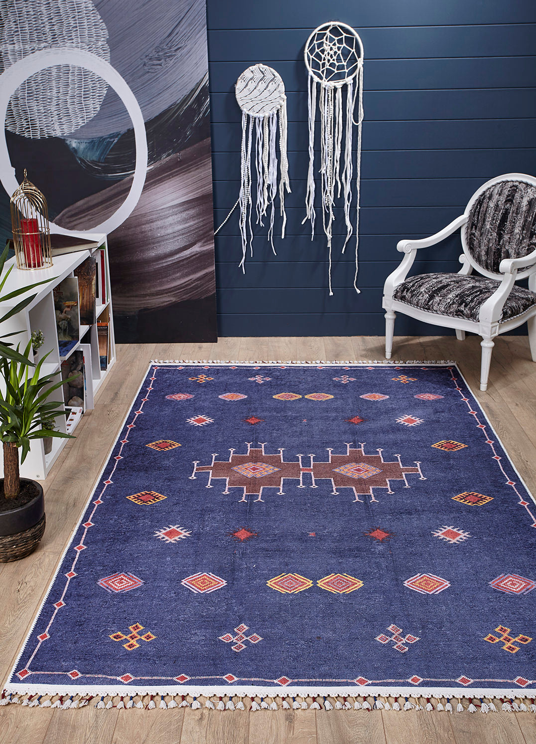 A stylish room features the Boho Chic Tapestry - Washable Rug - JR5083 in blue, paired with a white armchair, bookshelf, potted plant, and two white dreamcatchers on a dark blue wall over light wood floors.