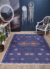 A stylish room features the Boho Chic Tapestry - Washable Rug - JR5083 in blue, paired with a white armchair, bookshelf, potted plant, and two white dreamcatchers on a dark blue wall over light wood floors.
