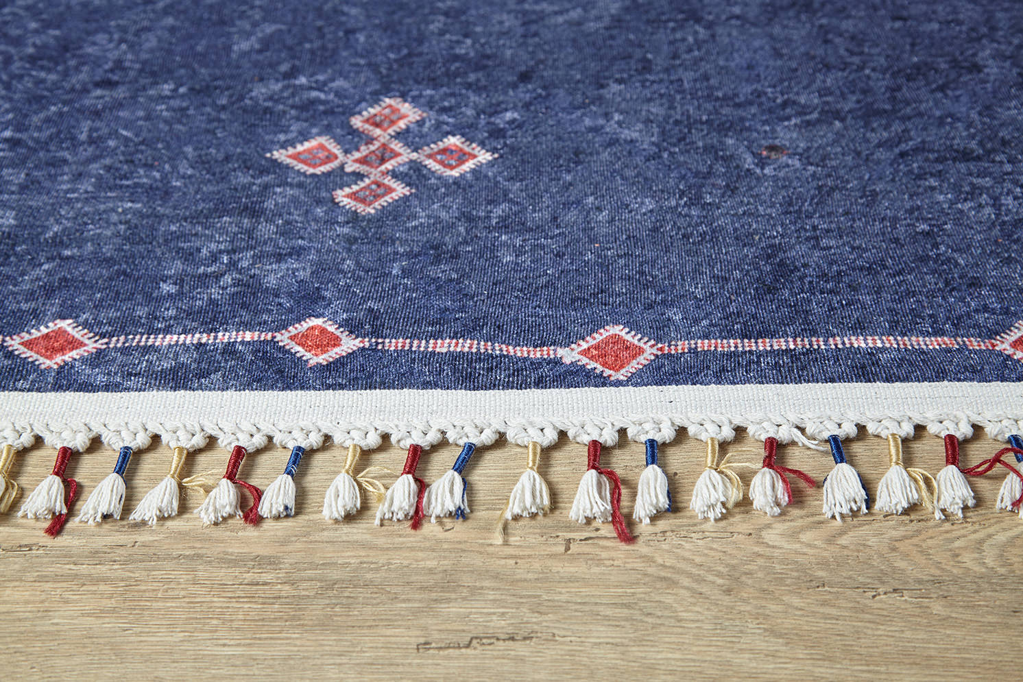 A close-up of the Boho Chic Tapestry - Washable Rug - JR5083, featuring blue with red and white geometric patterns and fringes woven with red and blue threads, displayed on a light wooden floor.