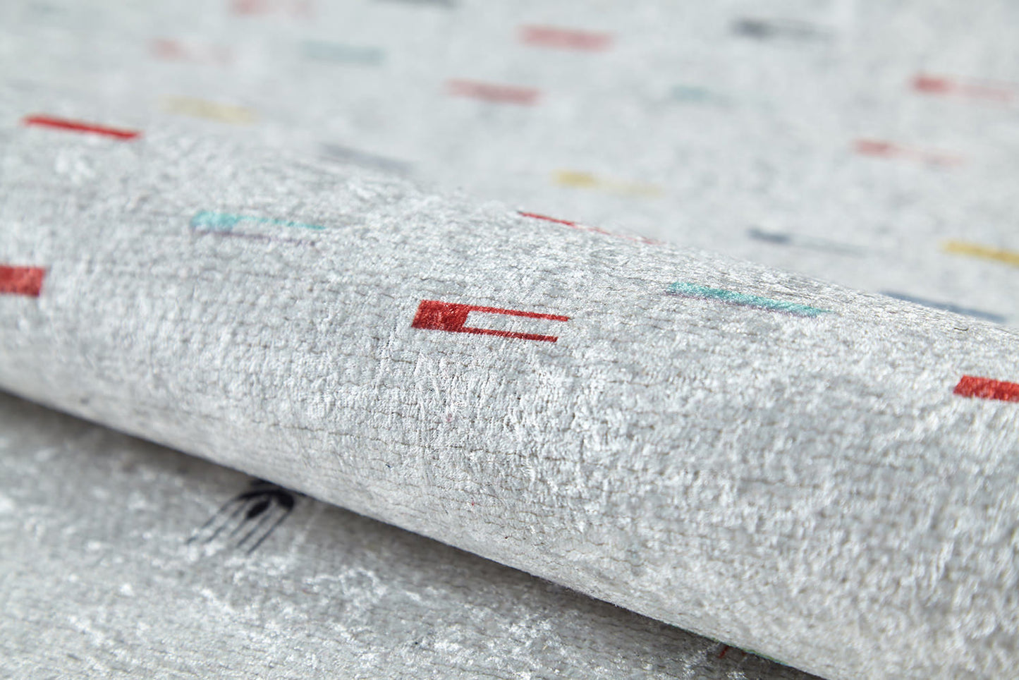 A close-up of the Boho Chic - Washable Rug - JR5103 reveals its textured, light gray surface with scattered red, teal, yellow, and black patterns. Slightly rolled to show detail—perfect for high-traffic areas.