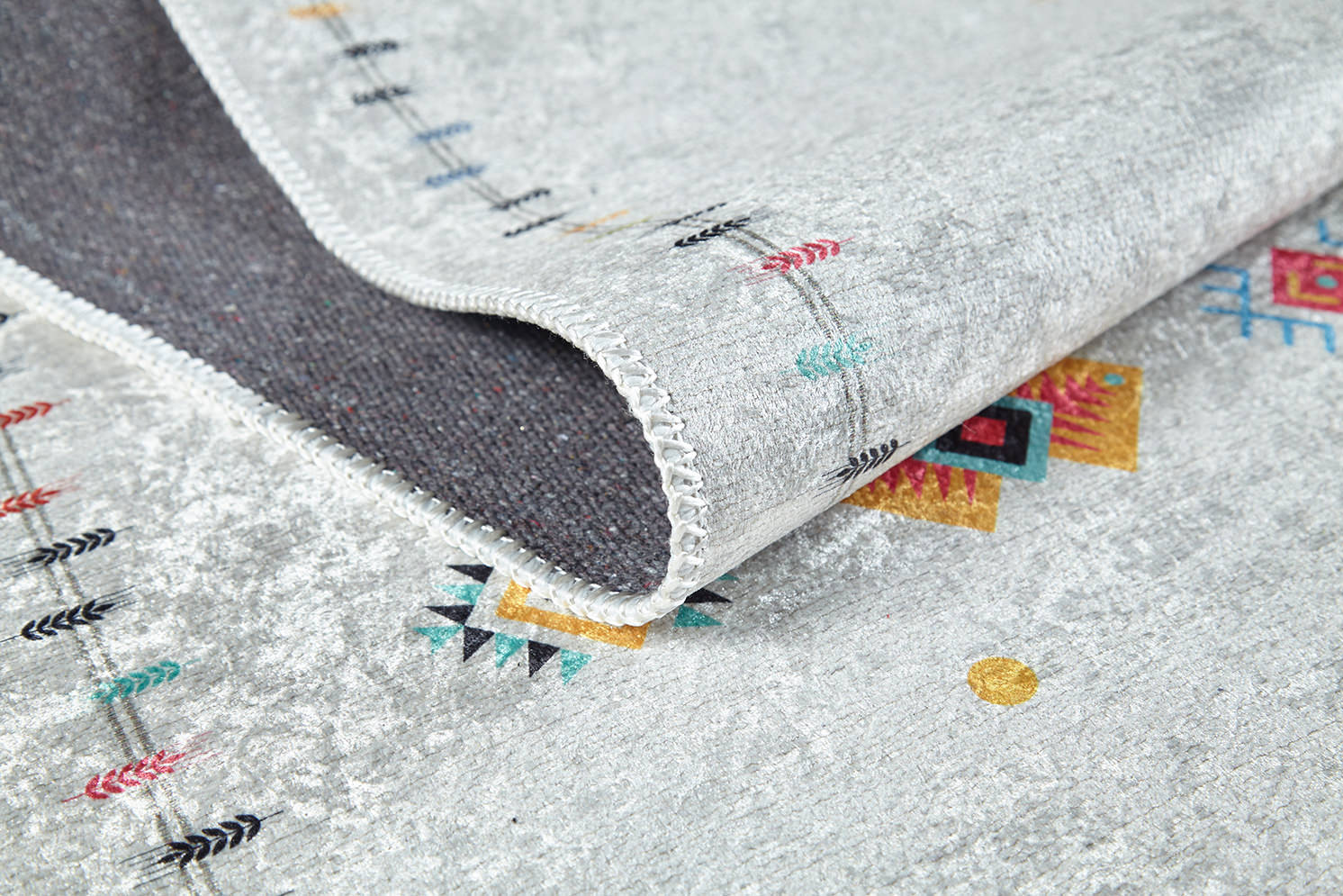 A close-up of the Boho Chic - Washable Rug - JR5103 in light gray, featuring colorful geometric patterns and black arrows. One corner is folded to show its dark underside and stitched edge—ideal for high-traffic areas.