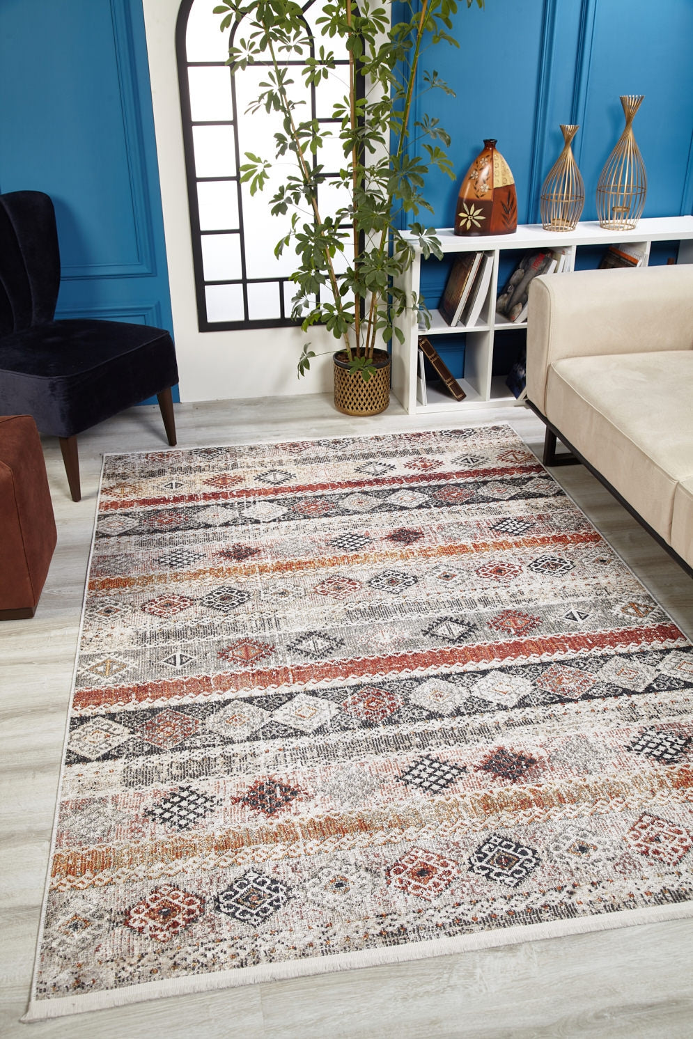 A modern living room featuring the Boho Oasis - Washable Rug - JR1618 in earth tones—ideal for high-traffic areas—paired with a beige sofa, tall leafy plant, decorative vases on a white shelf, black grille window, and blue paneled walls.