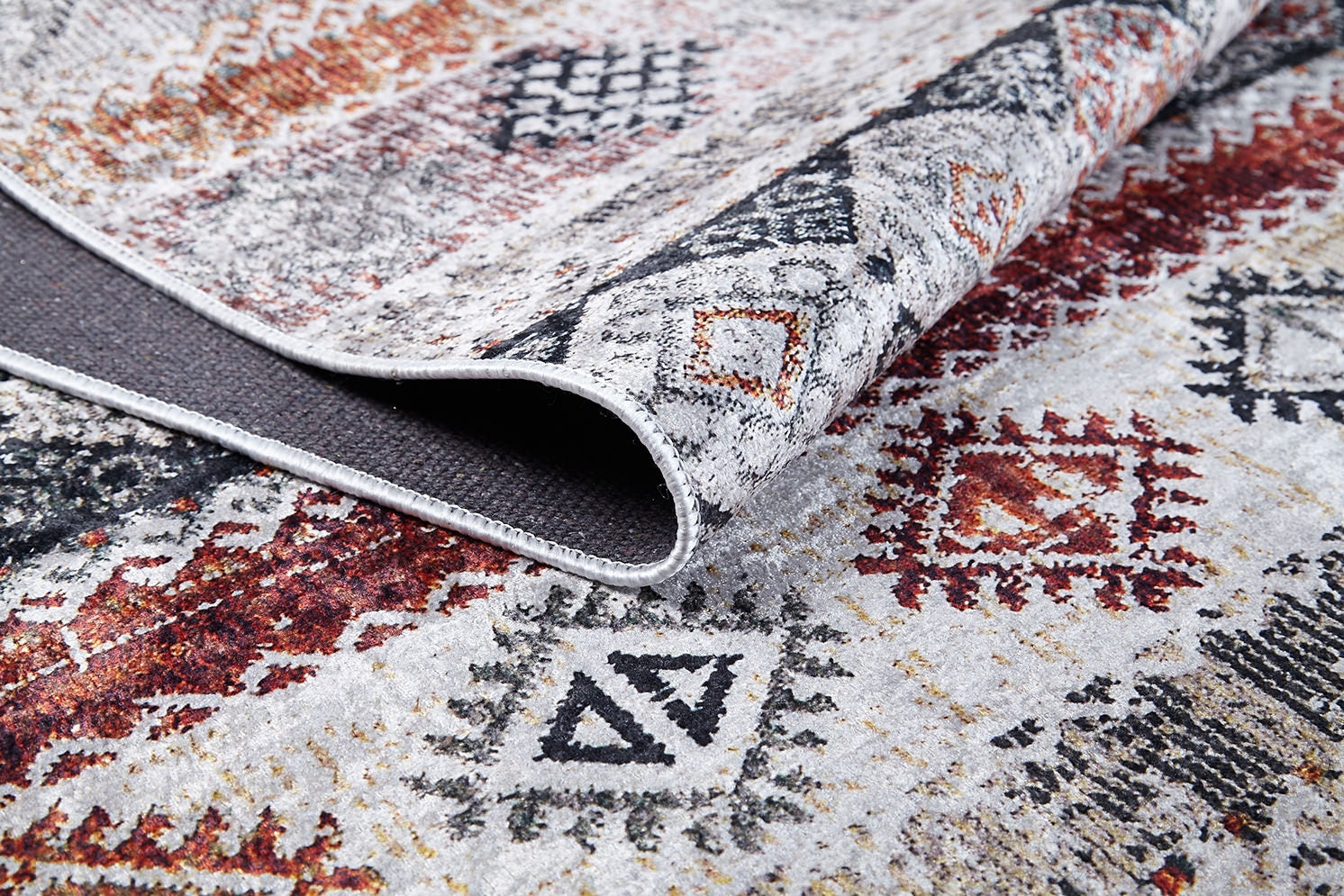 A close-up of the Boho Oasis - Washable Rug - JR1618, featuring bold geometric designs in black, red, and orange with a folded corner exposing its dark, textured underside—perfect for high-traffic areas.