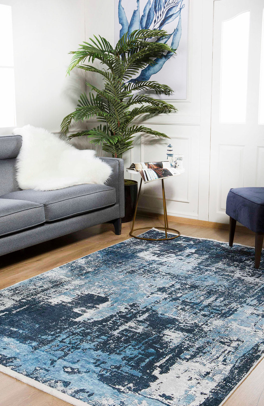 A modern living room features the Bold Blooms - Washable Rug - JR836 with blue patterns, a gray sofa with a white fur throw, large green plant, small side table with books, and blue ottoman—ideal for high-traffic areas with bright natural light.