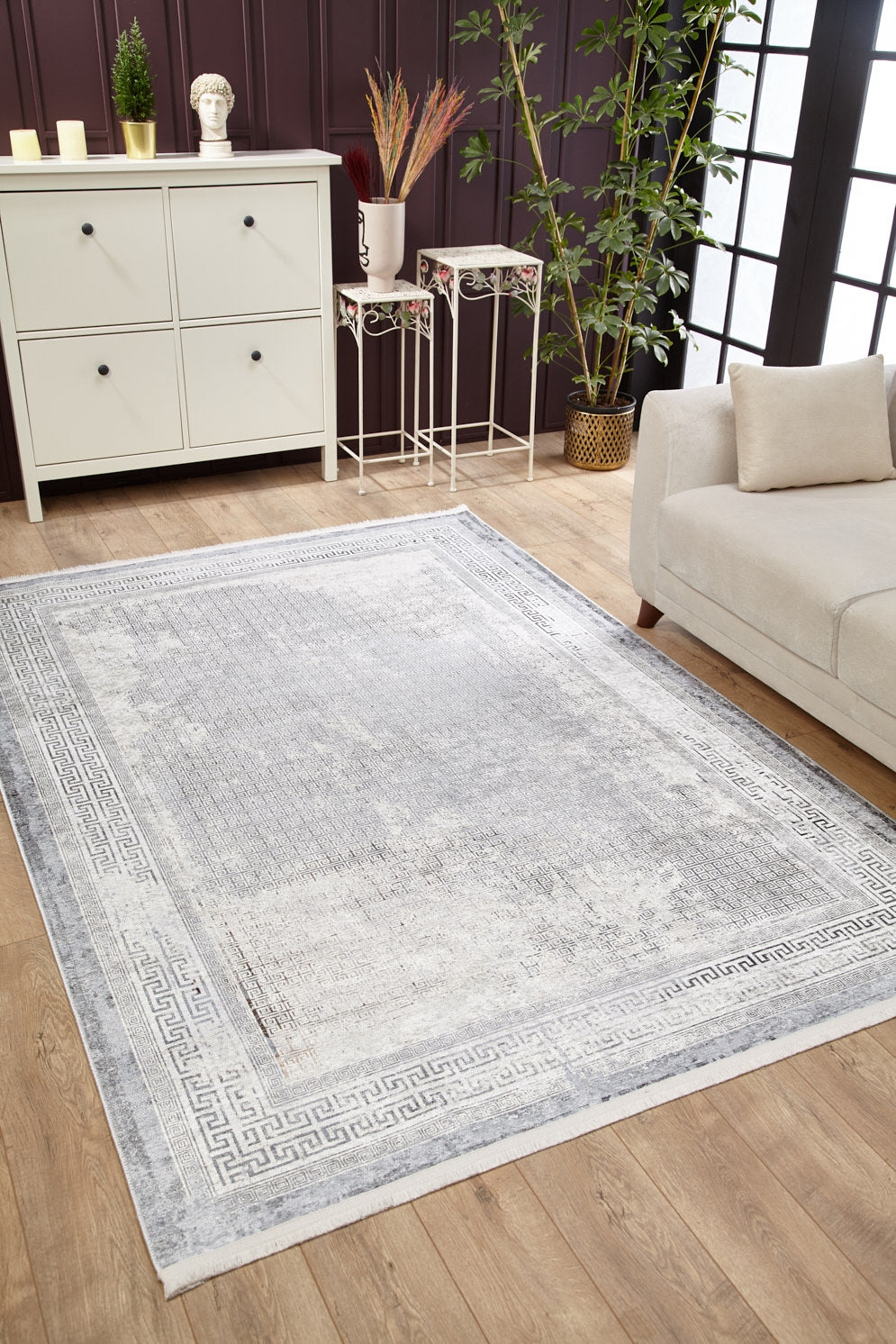 The Bold Borderline - Washable Rug - JR1984, in light gray-anthracite with a Greek key border, sits on a wooden floor in a modern living room with large windows, beige sofa, white cabinet, green plants, and decorative tables—perfect for high-traffic areas.