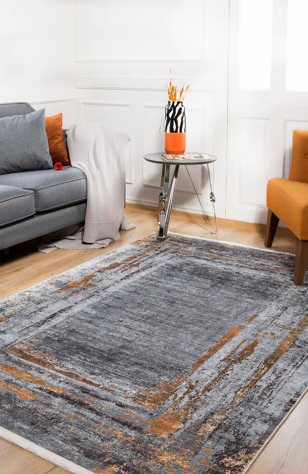 A modern living room featuring the Bold and Chic - Washable Rug - JR1198 in gray and orange, a matching sofa and cushions, cozy throw, round glass side table with vase, orange chair, and wood flooring—perfect for allergy-sensitive homes.