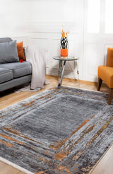 A modern living room featuring the Bold and Chic - Washable Rug - JR1198 in gray and orange, a matching sofa and cushions, cozy throw, round glass side table with vase, orange chair, and wood flooring—perfect for allergy-sensitive homes.