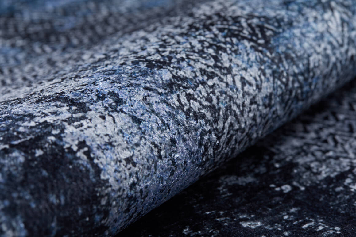 Close-up of the Bold and Minimalist - Washable Rug - JR1229, featuring a rolled-up textured surface with an abstract marbled pattern in blue, gray, and black—ideal for high-traffic areas seeking a stylish, weathered look.