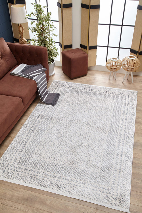 Load image 1 in gallery view. A modern living room with a brown sofa, gray and white striped blanket, the Borderline Brilliance washable rug in light gray, brown ottoman, two rattan side tables, potted plant, and large windows with bamboo shades.