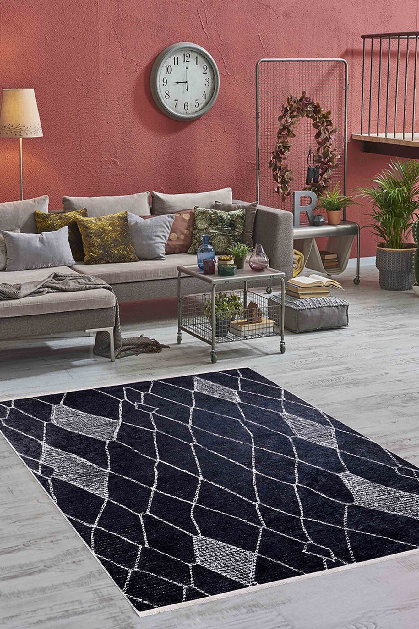 A modern living room with a gray sectional, assorted pillows, floor lamp, wall clock, potted plants, and the Byzantine Beauty - Washable Rug - JR1037 in black—ideal for high-traffic areas on light wood floors. The wall is muted red.