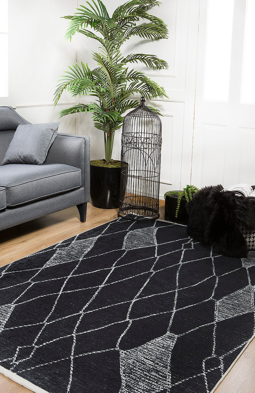 A modern living room features a gray sofa, a large leafy potted plant, black decorative birdcage, black pillows on a light wood floor, and the Byzantine Beauty - Washable Rug - JR1037—perfect for high-traffic areas with its stylish black and white pattern.
