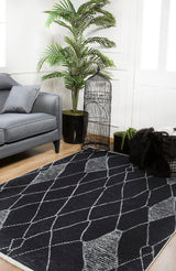 A modern living room features a gray sofa, a large leafy potted plant, black decorative birdcage, black pillows on a light wood floor, and the Byzantine Beauty - Washable Rug - JR1037—perfect for high-traffic areas with its stylish black and white pattern.
