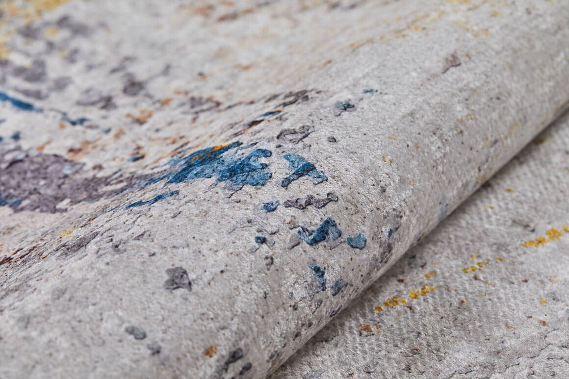 A close-up of Chromatic Kaleidoscope - Washable Rug - JR1483 reveals a textured, light gray surface with splashes of blue, yellow, and brown—an abstract design ideal for high-traffic areas.