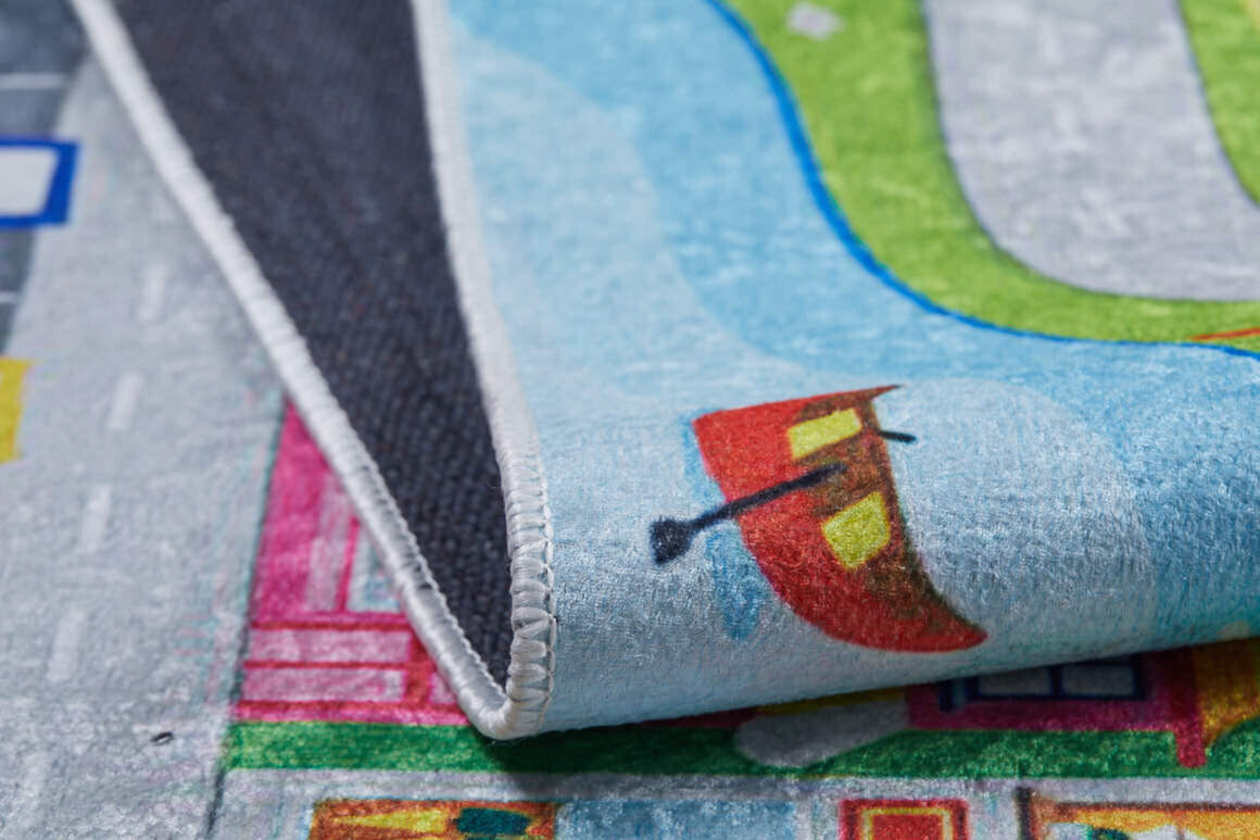 A close-up of the City Adventure - Machine-Washable Rug JRC039, featuring roads, grass, buildings, and a boat design; shown partially folded to display its black textured underside.
