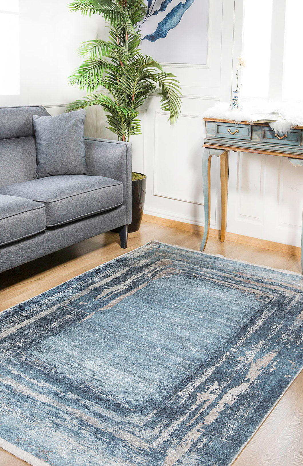 A bright living room features the Classic Bordered - Washable Rug - JR1197 in blue and gray, perfect for high-traffic areas, paired with a gray sofa, pillow, potted plant, blue desk with gold legs, and framed art under natural light.
