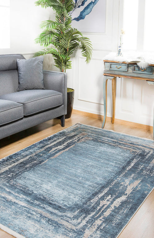 Load image 1 in gallery view. A bright living room features the Classic Bordered - Washable Rug - JR1197 in blue and gray, perfect for high-traffic areas, paired with a gray sofa, pillow, potted plant, blue desk with gold legs, and framed art under natural light.