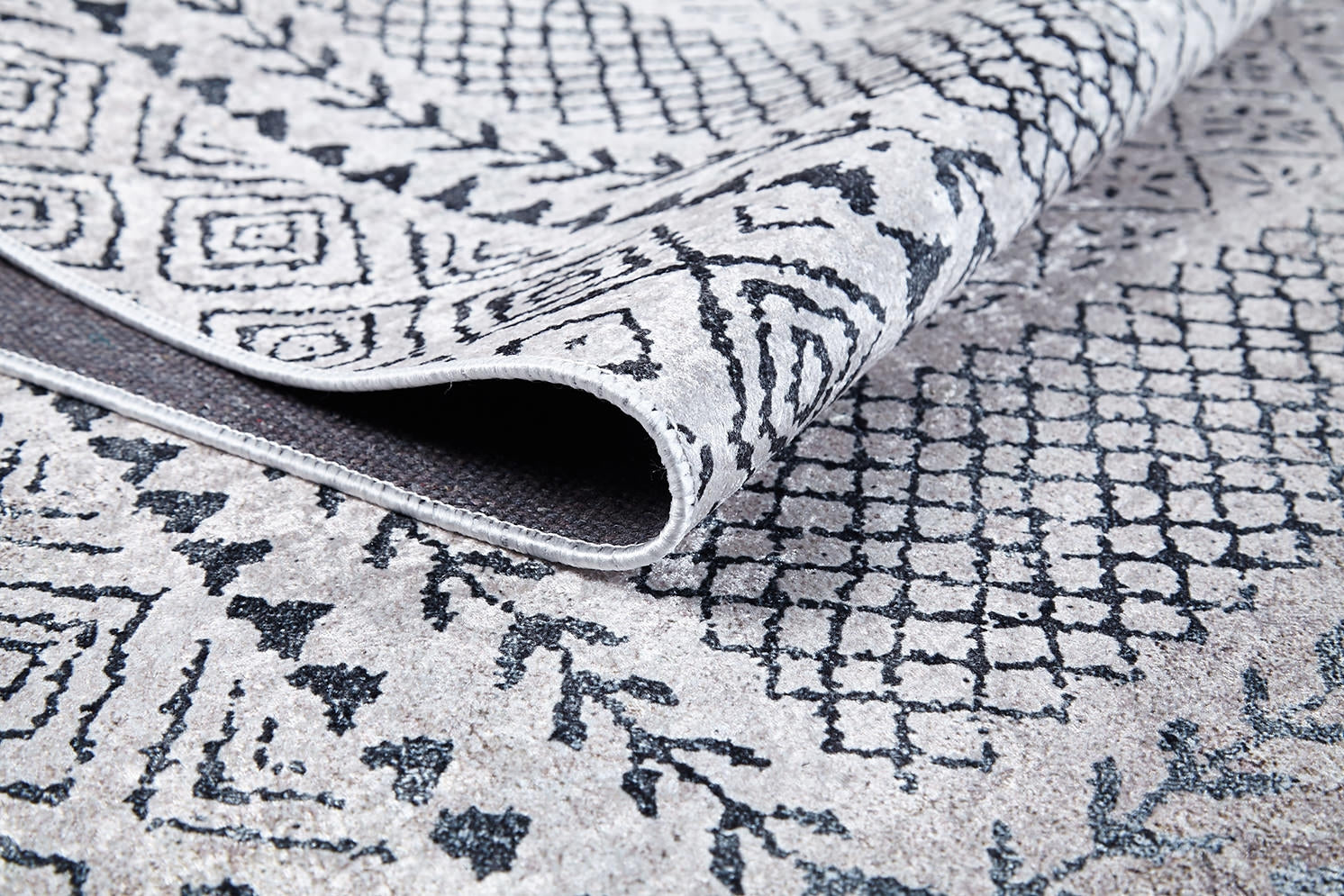 Close-up of a Classic Contrast - Washable Rug - JR1985 corner being lifted, showing geometric and abstract black patterns on a light gray background. The durable texture and weave of this gray-anthracite rug are clearly visible.