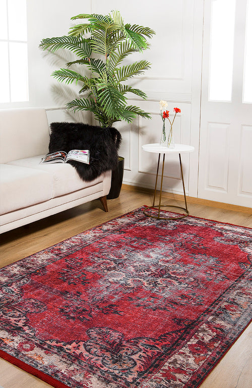 Load image 1 in gallery view. A cozy living room features the Classic Revival - Washable Rug - JR1374 in red, a cream sofa with a black pillow and magazine, a white round side table with flowers, and a potted plant by large white windows—ideal for high-traffic spaces.