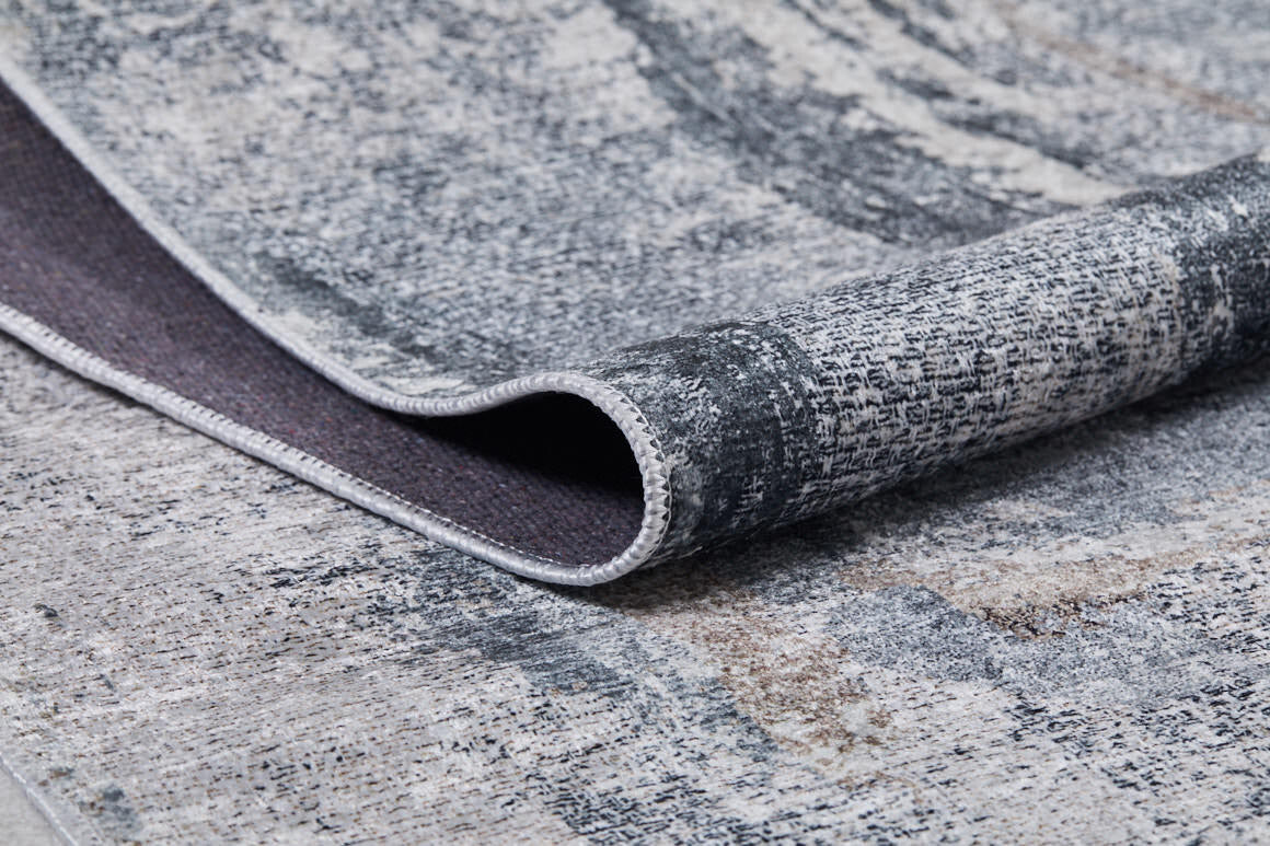A close-up of the Classic and Bold - Washable Rug - JR1529 in modern gray-anthracite with a textured, abstract pattern. One edge is folded to reveal the dark underside and stitched border—ideal for high-traffic areas.