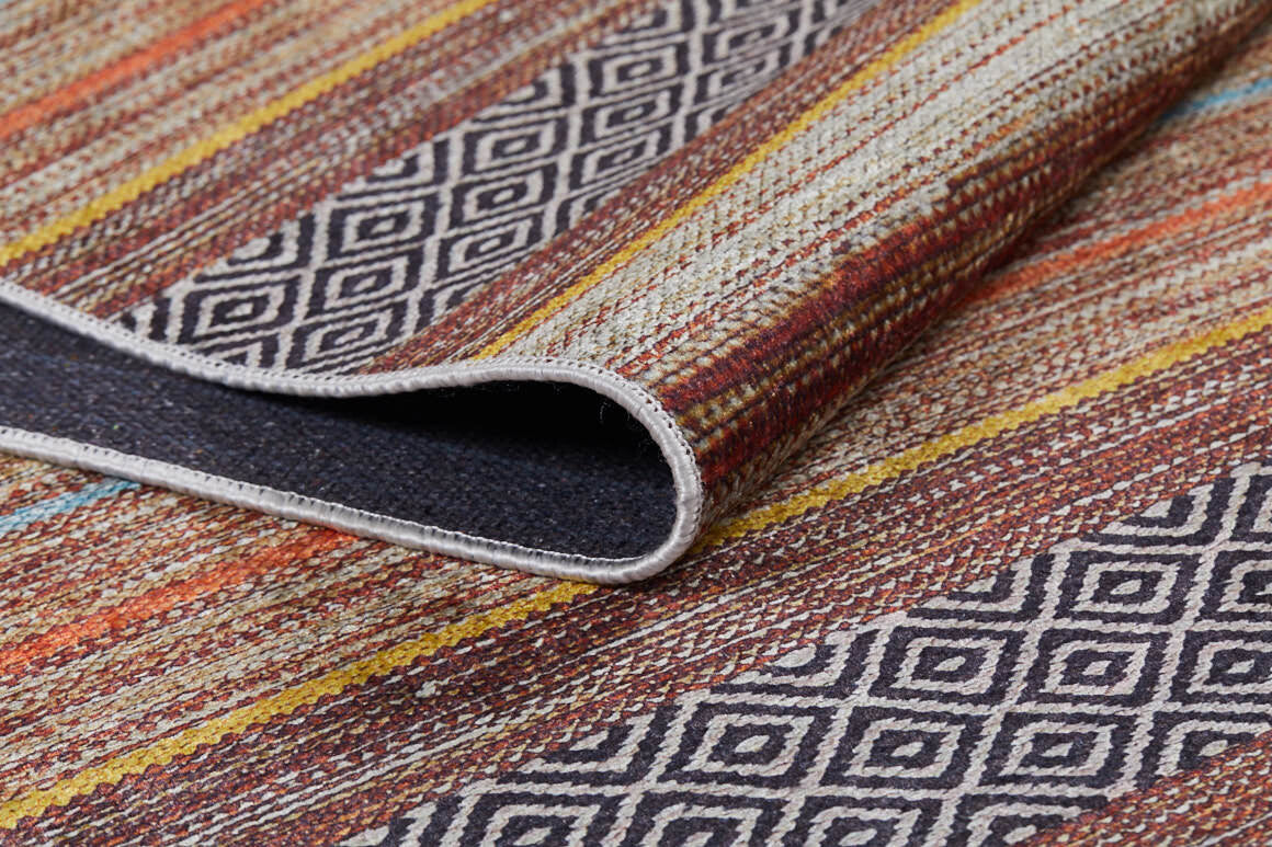 A close-up of the Coastal Breeze - Washable Rug (JR1549) showcases its geometric diamond pattern with colorful stripes. One corner is folded, revealing the allergy-sensitive rug’s underside.