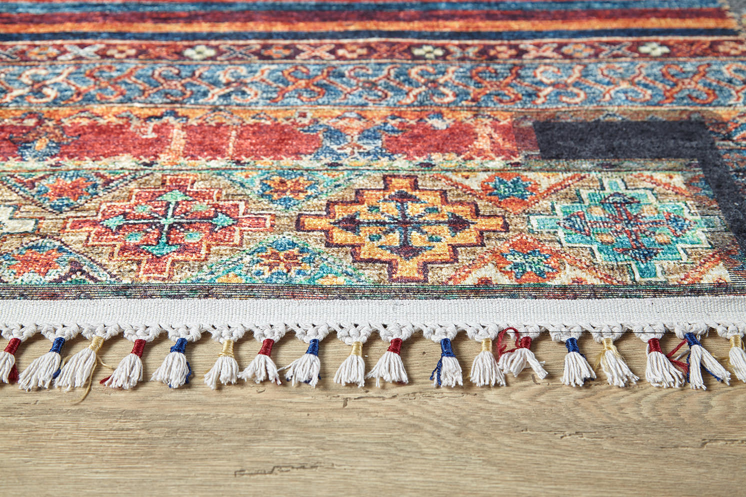 The Colorful Composition - Washable Rug - JR5107 features a vibrant multicolor geometric pattern in red, blue, gold, and teal with white fringe edged in blue and red threads, shown on a light wooden floor.