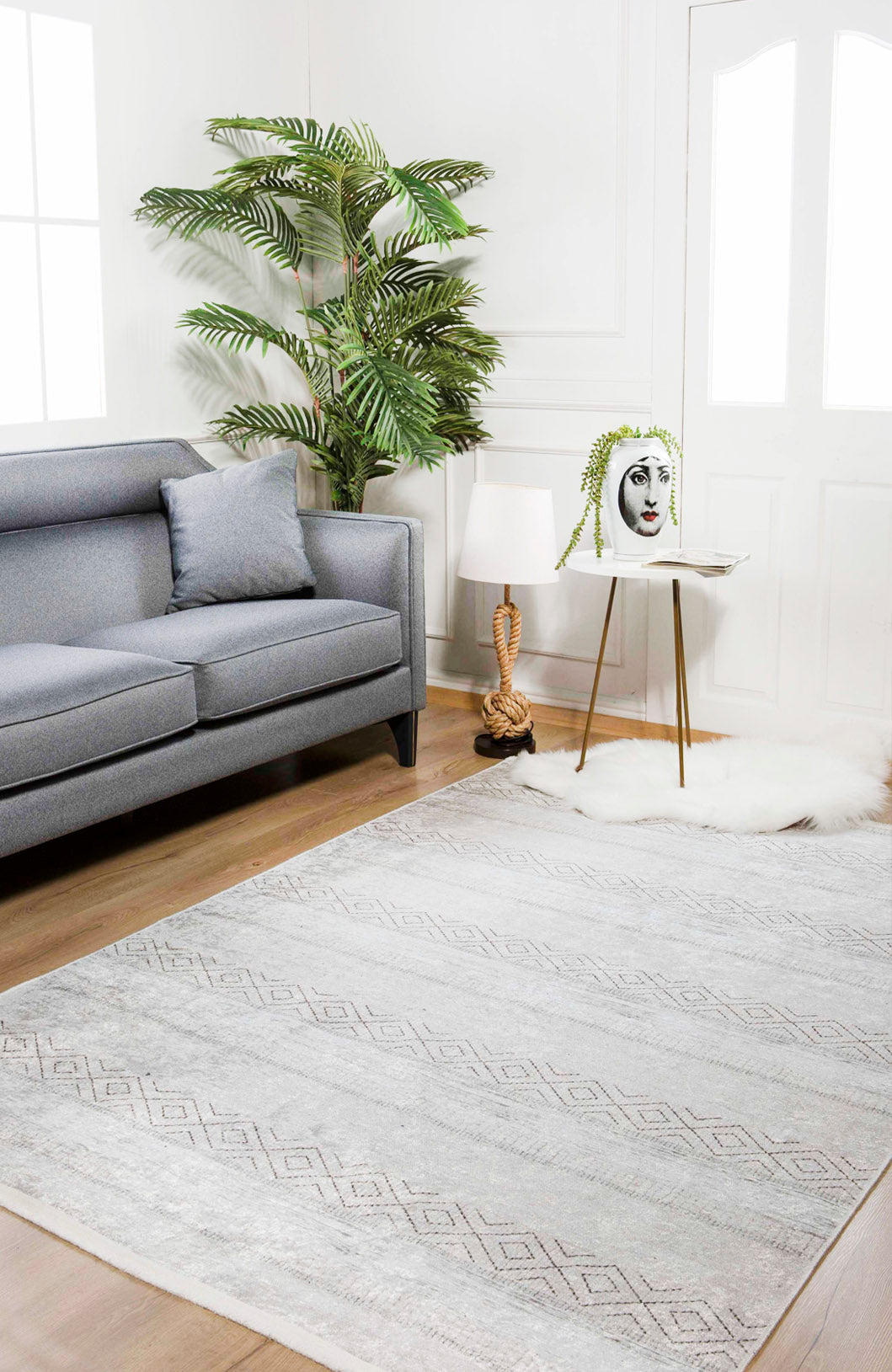 A modern living room with a gray sofa, cushion, tall green plant, face vase with trailing leaves on a table, decorative lamp, and the large Contemporary Coordinates - Washable Rug - JR750 in cream and beige on the wooden floor.