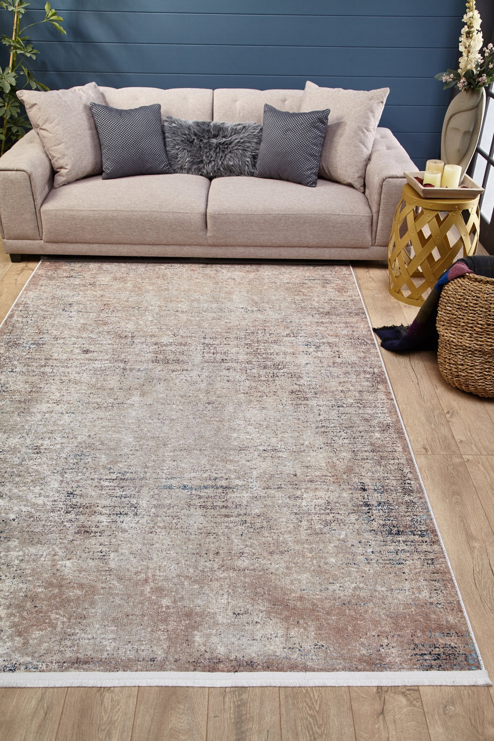 The Copper Glow - Washable Rug - JR1700, in cream and beige, sits on wood flooring by a light tan sofa with gray and beige pillows. Perfect for high-traffic areas, it’s styled with a round side table, candles, basket, and set against a blue wall.