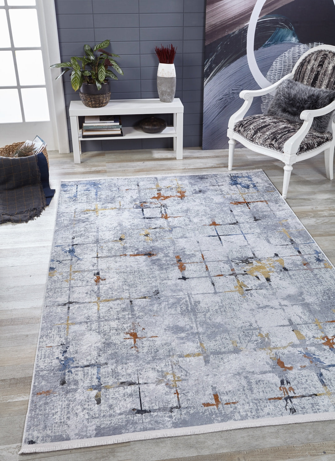 A modern living room features the Cross Country - Washable Rug - JR1753 in gray with an abstract pattern, a white cushioned chair, small white table with plants and decor, abstract painting, gray wall, and light wood flooring for a bright look.
