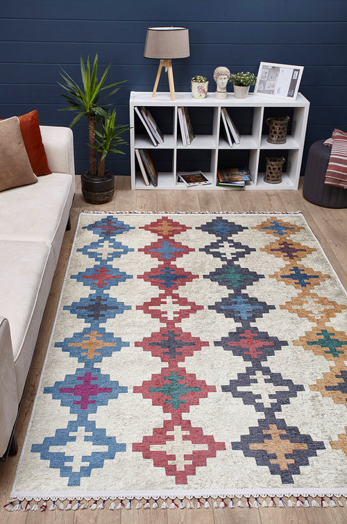 Load image 1 in gallery view. A living room features a beige sofa, patterned cushions, a Desert Mirage - Washable Rug - JR5033 with colorful geometric design, a white shelf holding books and decor, a potted plant, and a table lamp set against dark blue walls and wooden flooring.