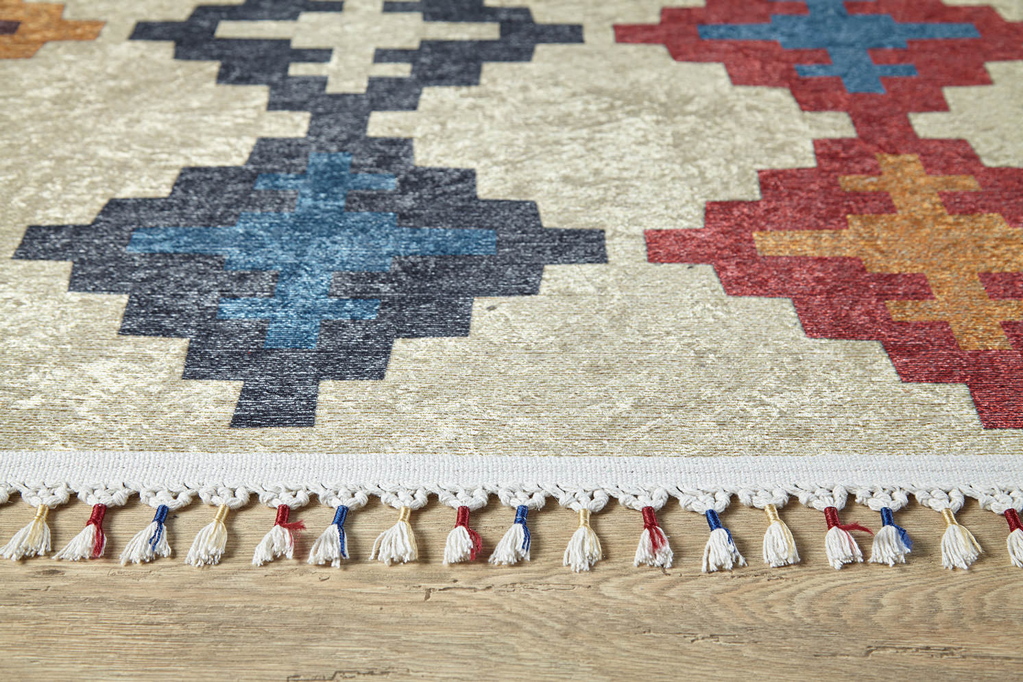 A close-up of the Desert Mirage - Washable Rug - JR5033, a flat weave with blue, navy, red, and orange geometric patterns on beige. The edge features white tassels with red and blue accents. This allergy-friendly rug sits on a wooden floor.