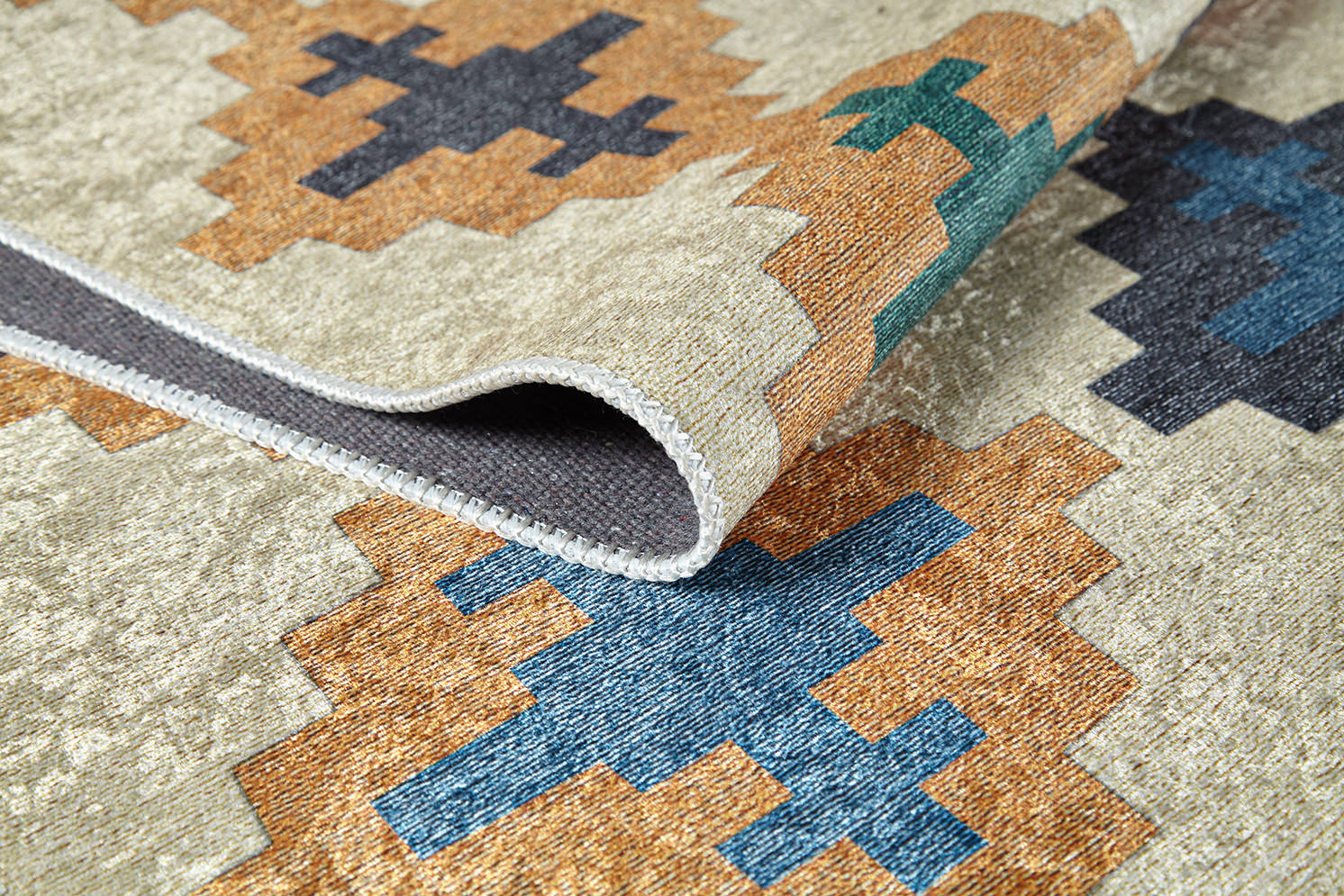 A close-up of the Desert Mirage - Washable Rug (JR5033), a flat weave with geometric shapes in blue, green, brown, and black on beige. The washable rug is slightly folded to display its edge and thickness.