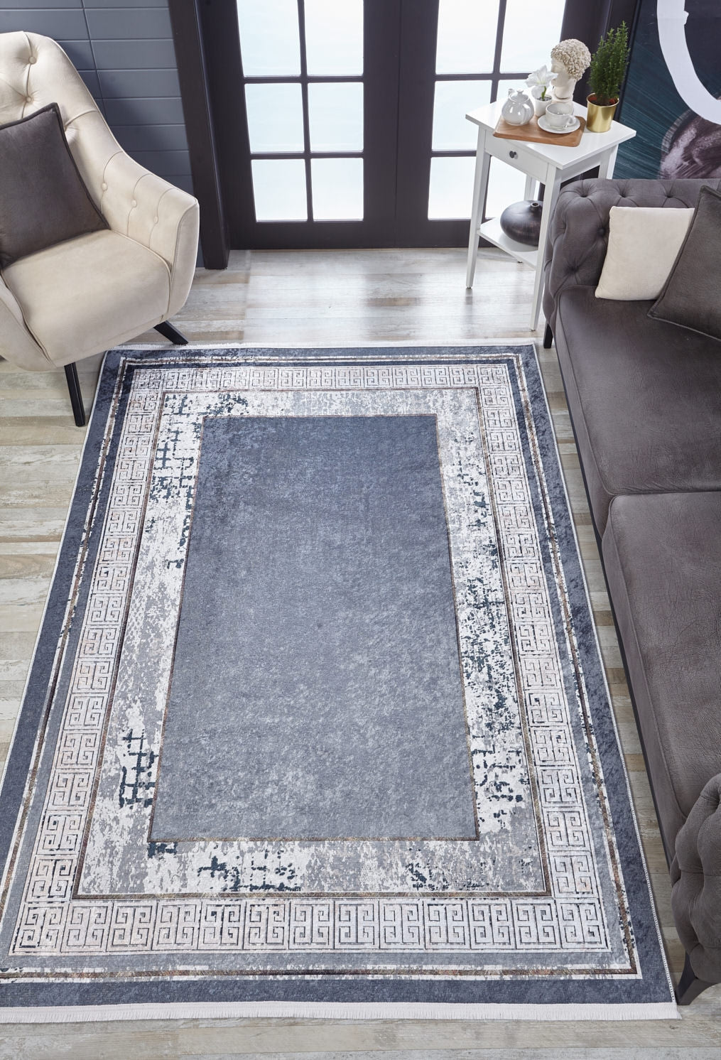 A modern living room features the Dramatic Boundaries - Washable Rug - JR1810 in gray, a beige armchair, gray sofa, white side table with potted plant, wood floor, and double glass doors—perfect for high-traffic spaces.