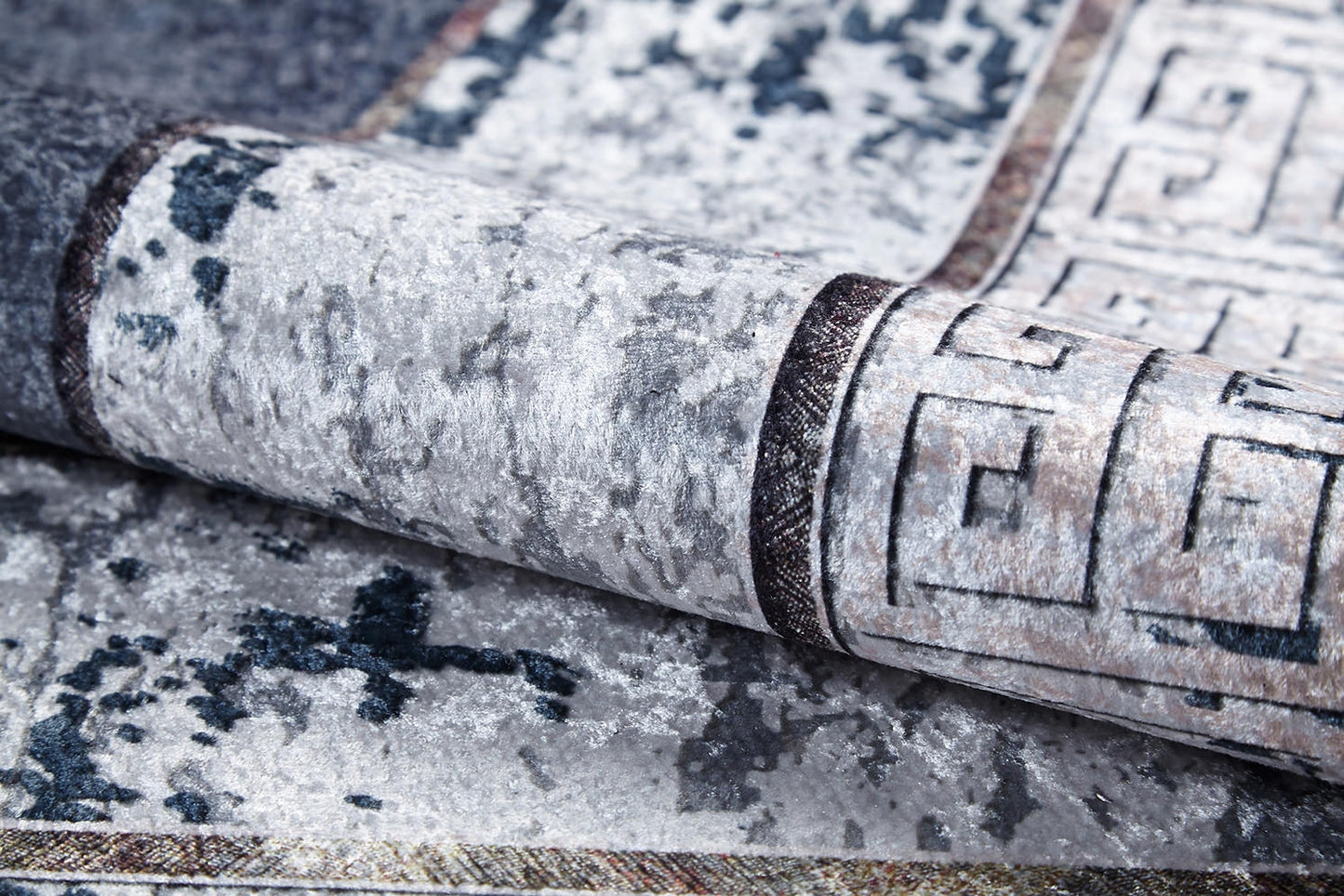 A close-up of the Dramatic Boundaries - Washable Rug (JR1810), folded to show its gray, textured, mottled pattern and geometric border in dark and light tones—perfect for high-traffic areas.